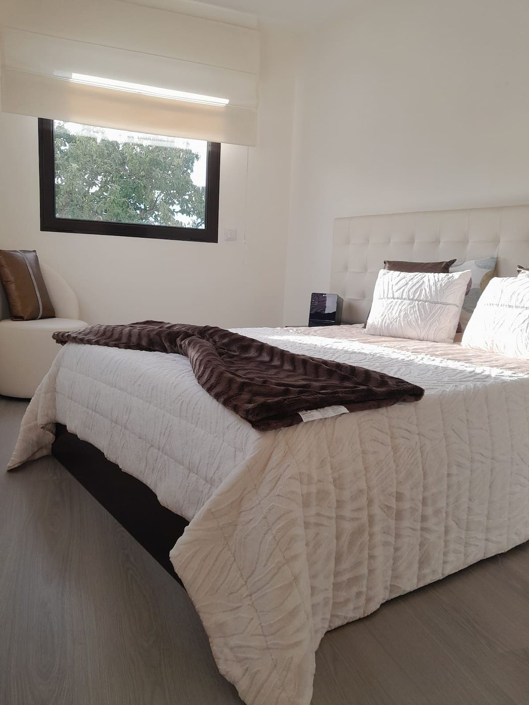 Quarto de casal com cabeceira estofada, roupa de cama branca e manta castanha, duas janelas